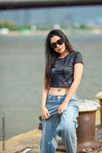 Stylish young Asian woman in sunglasses, black crop top and baggy blue jeans posing on a riverside dock
