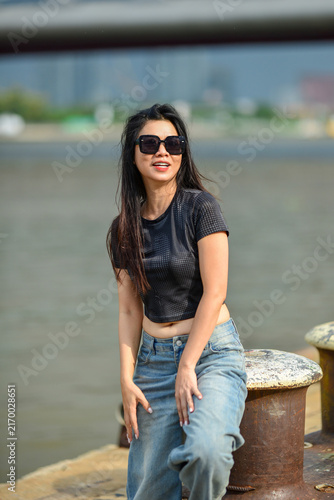 Confident young Asian woman in black crop top and sunglasses posing by a riverside dock in sunlight