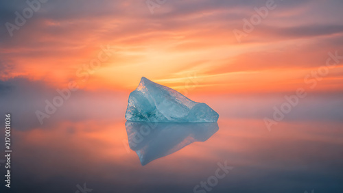 small iceberg floating on calm water with soft reflection under glowing orange sunset sky and fog