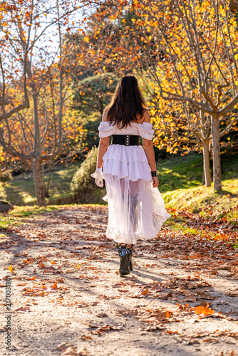 Woman wearing a white gothic dress, black corset and boots walking along a forest path surrounded by autumn foliage and golden leaves