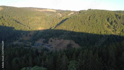 Smooth aerial view over lush green countryside with open fields, dense trees, winding rural roads and peaceful natural scenery in bright daylight.
