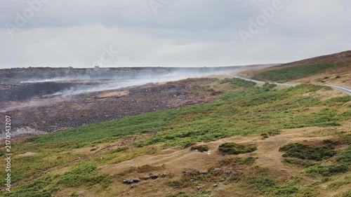 Smoke drifting across scorched earth after moorland fire, blackened ground beside green hillside and empty road, wide shot