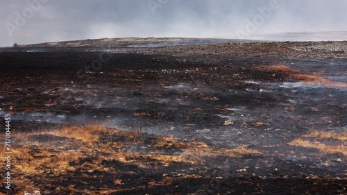 Burnt moorland landscape following grass fire, smoke rising from blackened earth and dry grass under overcast sky, wide view