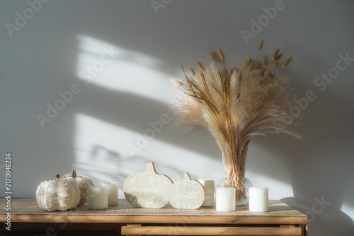 Cozy warm autumn home interior - white knitted pumpkins, wooden statuette, candles, dried flowers on the wooden console with white wall background. Scandinavian minimalist hygge home fall decor.