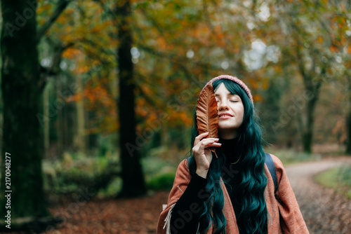 Portrait of Young smiling woman in hat partially covering her face with striking red fallen leaf enjoying autumn walk in forest. Feeling harmony, reunion with nature. Relaxing, personal fulfilment