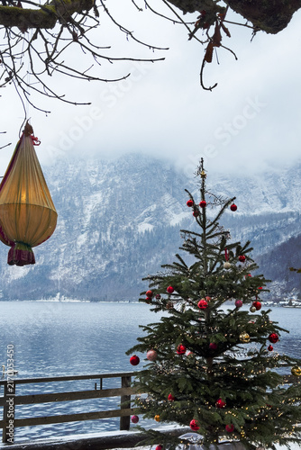 Vertical shot of decorated outdoor Christmas tree with red baubles on lake shore in Hallstatt, Austria. Snow covered mountains and mist in winter holiday landscape.