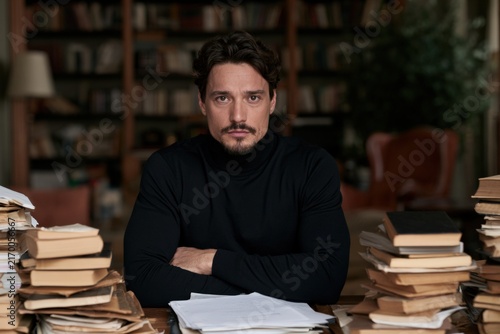 Man sitting at desk surrounded by stacks of books and papers indoors