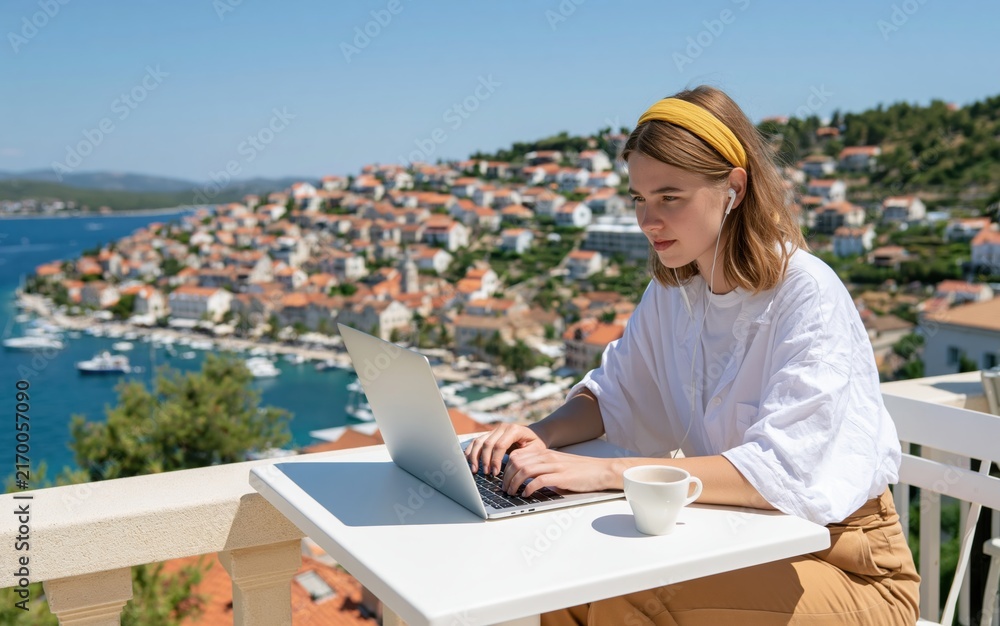 Fototapeta premium Woman working on laptop at a sunny coastal terrace in a beautiful mediterranean town