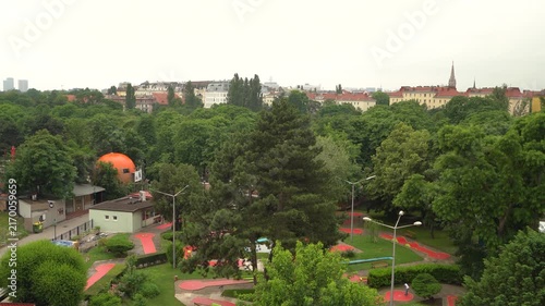 Aerial video of Vienna's Prater Park playground with colorful recreational equipment, lush green trees, and historic city architecture in background on overcast day.