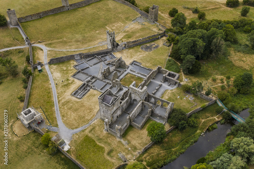 Aerial view of Kells Priory, a sprawling medieval monastic site with stone ruins, defensive walls, and lush green fields situated along a river in County Kilkenny, Ireland.