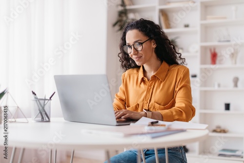 Focused Asian Woman Working On Laptop At Home Office, Smiling While Typing On Computer, Sitting At Desk With Office Supplies. Engaged In Professional Work And Productivity. Business Concept.