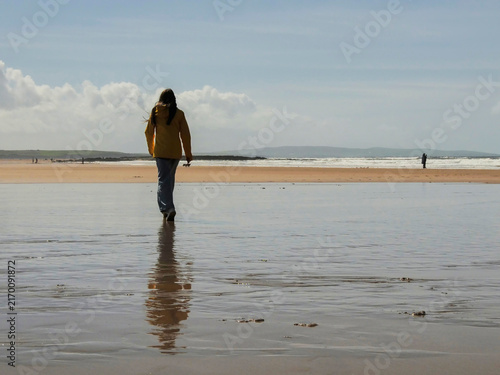 Obraz na plátně Person walks alone on wet beach, reflecting in shallow water