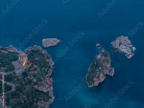 Aerial view of the historic Torre Bennistra perched on a cliff overlooking the Tyrrhenian Sea with rocky outcrops and deep blue water in Castellammare del Golfo, Sicily, Italy.