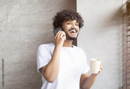 фотографія Joyful South Asian Man In His 20s Smiling And Engaging In A Phone Conversation While Holding A Coffee Cup, Relaxing At Home In A Cozy Environment