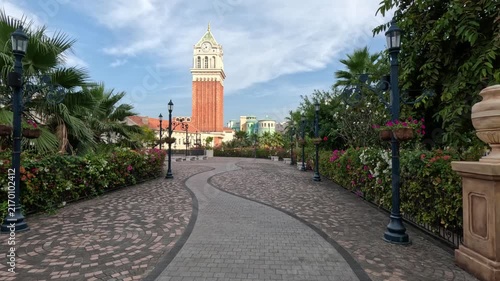 Wide shot of a beautiful alley in a Vietnamese park featuring a mosaic-paved path, manicured flower bushes, palm trees, and a large clock tower in the background.