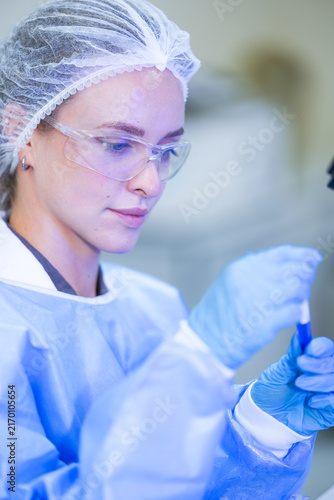 Fotografía A woman researcher in white laboratory clothing, safety glasses and blue gloves rolling the blood tube between gloved fingers inside a university biotechnology laboratory