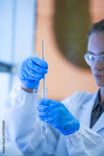 Bild auf Leinwand A young female lab scientist in white laboratory clothing, safety glasses and blue gloves holding tube and rod at the hood ledge in a modern medical research laboratory