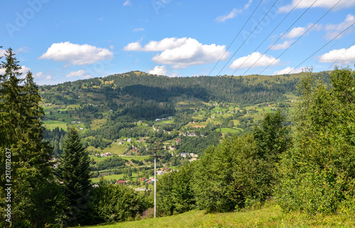 Panoramic view of a mountain valley with a small village nestled among forests and green meadows under a bright blue sky with scattered clouds. Carpathian Mountains, Ukraine