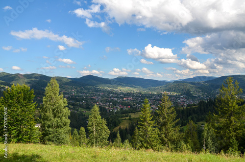 Panoramic view of a mountain valley with a small village nestled among forests and green meadows under a bright blue sky with scattered clouds. Carpathian Mountains, Ukraine