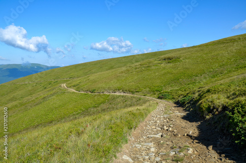 Rocky mountain trail winds across rolling green hills under a bright blue sky with scattered clouds, evoking peaceful hiking, outdoor adventure, wide-open landscapes and summertime exploration