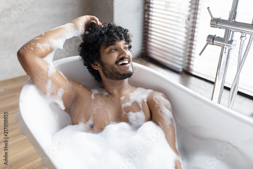 Fotografie Smiling Young Man With Curly Hair Relaxing In Bathtub Filled With Foam, Showing Joy And Contentment, Enjoying Personal Time In Luxurious Bathroom