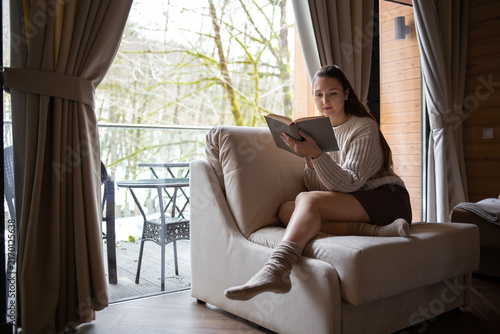 A young woman sitting in an armchair by a large window in a country house, reading a book, wearing a warm knitted beige sweater, brown skirt and cozy socks, with a snowy winter forest visible outside.