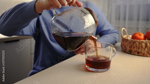 A woman pours a dark liquid from a glass pitcher into a transparent cup. The table is adorned with a woven basket filled with oranges, adding warmth to the scene.