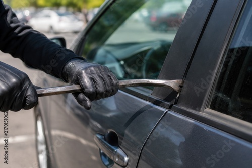 A criminal wearing black leather gloves uses a metal crowbar to pry open a locked car door window.