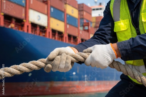 A dock worker wears gloves while pulling a heavy mooring rope on a cargo ship at a busy port terminal.