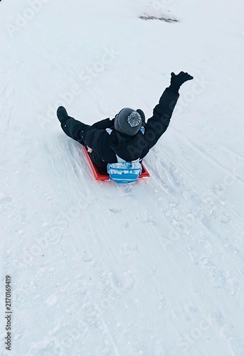 Foto Happy person riding red snow sled toboggan down snowy hill with arm raised in wi