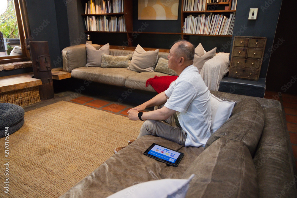 Obraz premium Senior Asian man sitting on suede sofa in living room using tablet near bookshelves