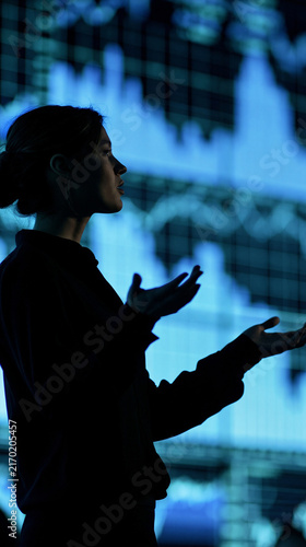 Analyzing Financial Trends: A focused businesswoman gestures towards a dynamic blue digital graph, symbolizing data analysis and financial market insights.