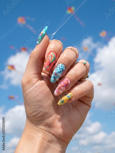 A hand with beautifully decorated nails adorned with vibrant floral patterns and a checkered design, set against a backdrop of a kite festival in the sky