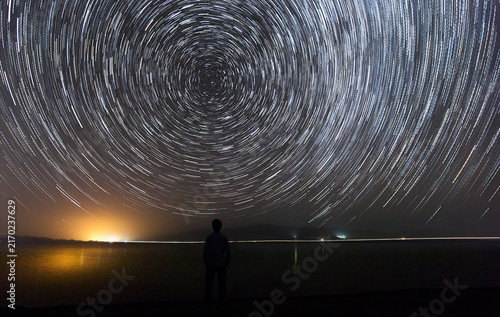 Cuadro en lienzo A long-exposure astrophotography shot shows circular star trails in the night sky, with a silhouetted figure standing by calm water reflecting distant light