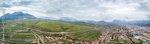 Canvastavla A high-angle panoramic view of a town nestled among green hills, fields, and distant mountains under a cloudy sky