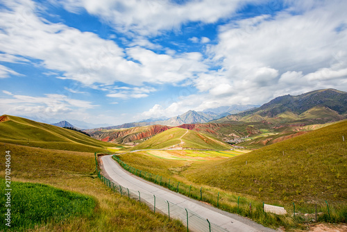Wall Mural A winding paved road cuts through rolling grassy hills and colorful mountain valleys under a bright blue sky with scattered white clouds