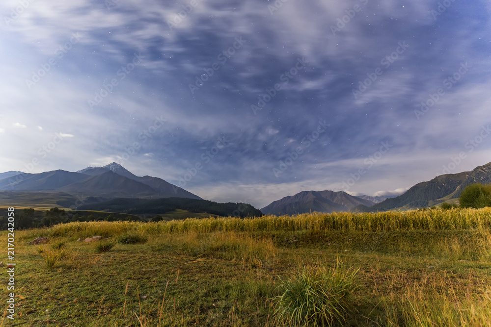 Obraz premium A wide landscape of grassland stretching toward distant mountain peaks under a blue sky with thin clouds, capturing a serene natural outdoor scene.