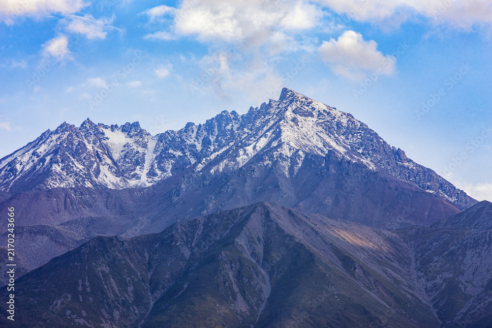 Obraz premium A rugged mountain range with snow-capped peaks stands beneath a bright blue sky dotted with white clouds, showcasing natural alpine scenery.