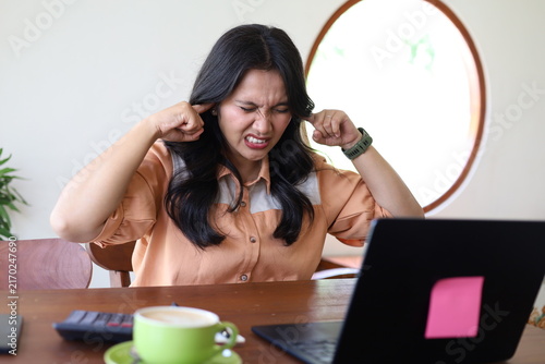Konstfotografi A portrait of a woman looking annoyed and exasperated as she covers her ears with her fingers while working remotely on a laptop in a coffee shop