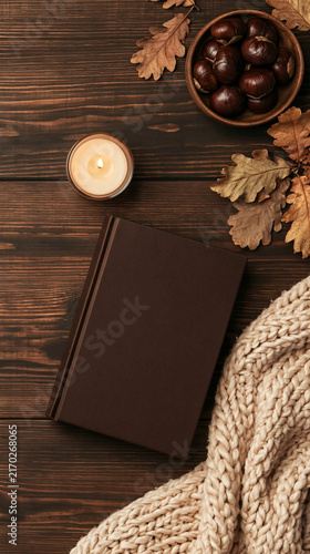 Autumnal Comfort: A warm candle glows beside a closed book and cozy knitted blanket, surrounded by fallen oak leaves and a bowl of chestnuts, evoking a sense of peaceful autumn repose and hygge.