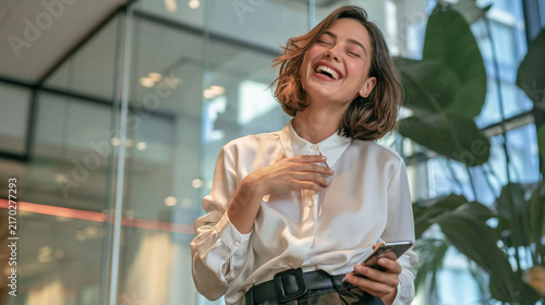 Joyful Woman Laughing: A young woman with a radiant smile and eyes closed in mirth, holding a smartphone, expresses pure happiness in a modern office environment.