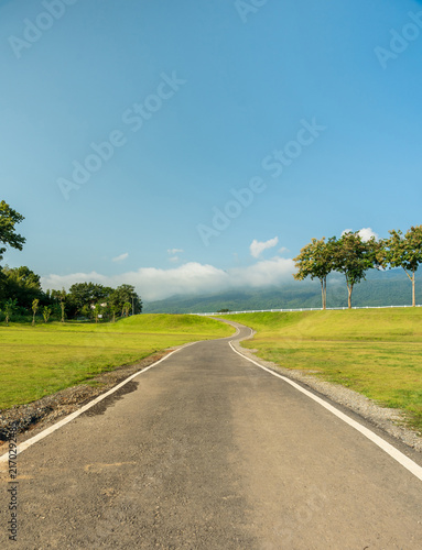 Winding country road curving through a green field toward misty mountains under a bright blue sky with soft clouds.