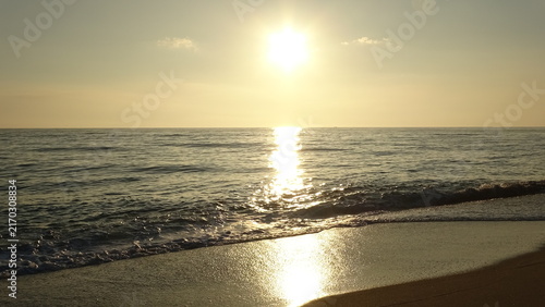 фотографія The sun has risen above the horizon, appearing brightly in the upper center, with its light reflecting down onto the restless sea and small swells rolling towards the beach