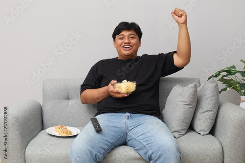 Quadro su tela A heavyset man sitting on the sofa watching TV raises a clenched fist to celebrate his favorite soccer team's victory, while a bowl of chips and a doughnut sit beside him