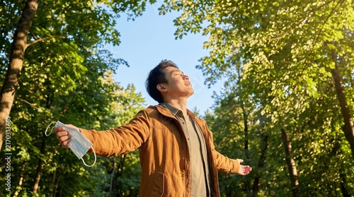 Young Asian man breathing deeply beneath a blue sky in a green park while holding a face mask, expressing freedom, hope and awareness of clean air and healthy environmental quality.