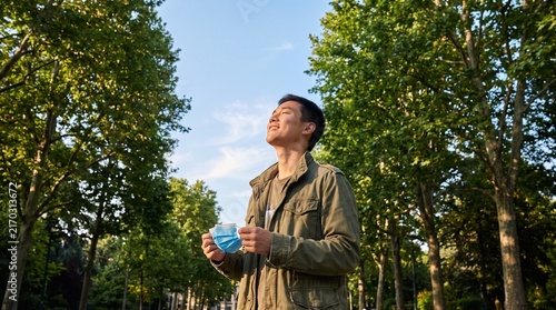 Young Asian man breathing deeply beneath a blue sky in a green park while holding a face mask, expressing freedom, hope and awareness of clean air and healthy environmental quality.