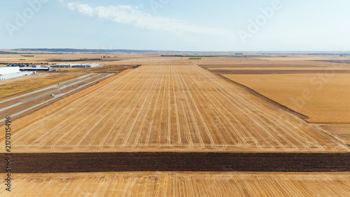 Aerial view of harvested agricultural fields with stubble patterns and a dark strip of plowed soil, with industrial buildings in the distance in Indjija, Vojvodina, Serbia.