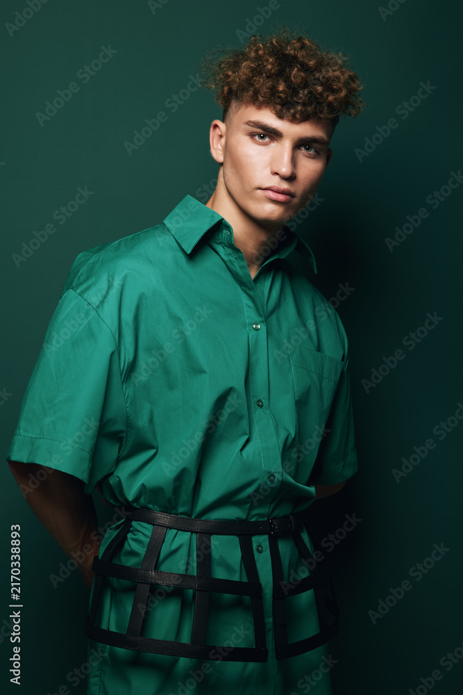 Naklejka premium Young man with curly hair wearing green shirt and black striped harness standing against green background in studio. Fashion portrait and casual pose with serious facial expression and male model.
