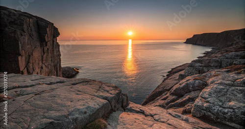 Bild auf Leinwand Rocky coastal cliff landscape with bright sun star reflection glowing on calm ocean horizon at sunset