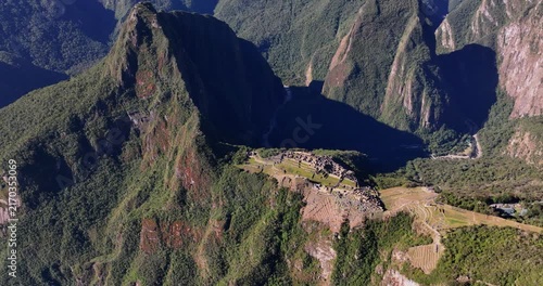 Aerial high angle view of the Historic Sanctuary of Machu Picchu nestled among lush green Andean mountains and steep terraced slopes under bright sunlight Peru
